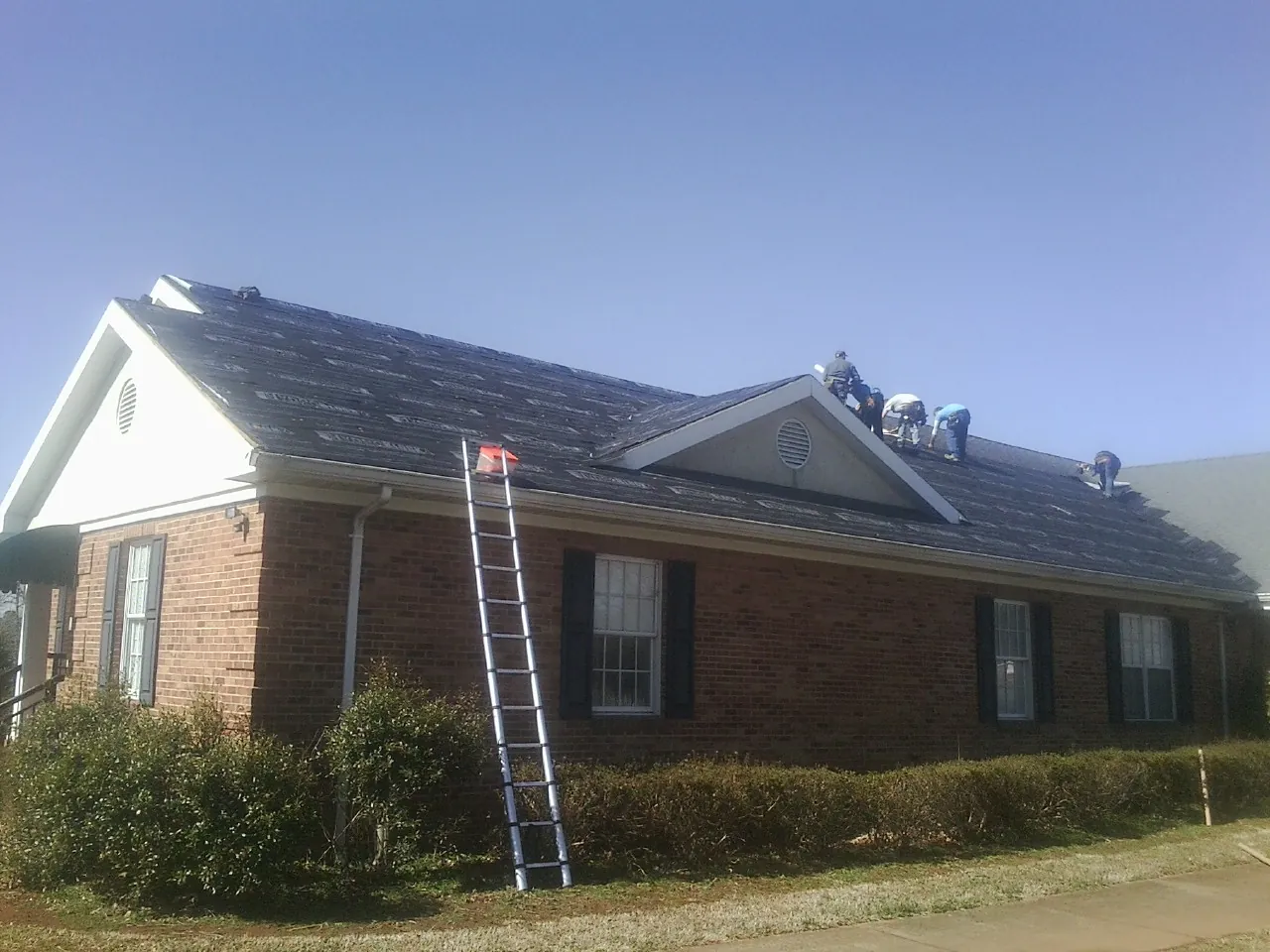 Crew tearing shingles off a brick ranch roof with a ladder set — Metal Roof Repair work by our crew in Runnells
