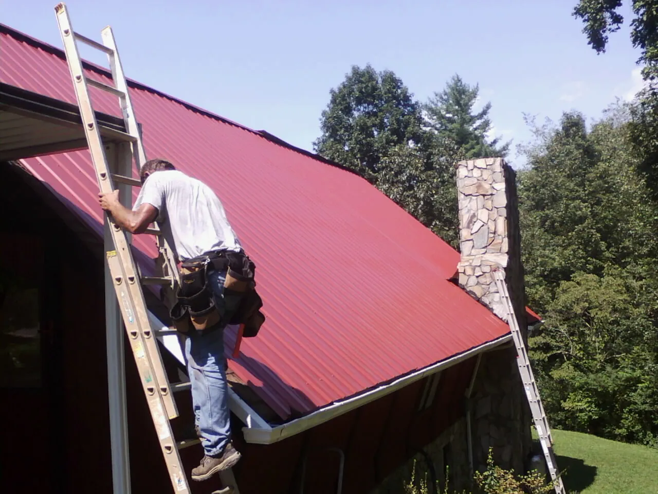 Roofer on a ladder beside a red metal roof and stone chimney — Soffit & Fascia Repair by Roofing Brothers in Runnells